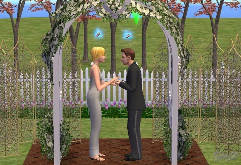 Screenshot of The Sims 2 showing a male and female Sim exchanging rings under a floral wedding archway outdoors. The female Sim has blonde hair and wears a long white wedding dress, while the male Sim wears a classic black tuxedo. They are standing on a brick path surrounded by a white picket fence and trees.