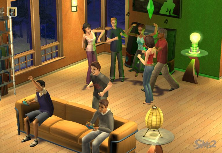 Screenshot of The Sims 2 displaying an isometric view of a lively teen house party in a living room at night. In the foreground, two male Sims are sitting on a bright orange sofa playing video games with controllers. In the background, a group of Sims is dancing together in front of a stereo system. The room features wood floors, large windows showing a dark, tree-lined exterior, and the classic Sims 2 logo is visible in the bottom right corner.